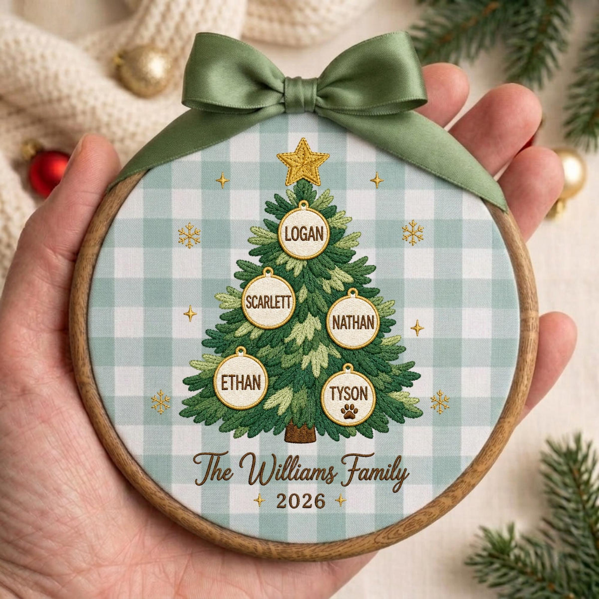 May include: A round wooden embroidery hoop ornament featuring a light green and white buffalo check fabric background. An embroidered green Christmas tree is centered with a gold star, five circular ornaments with names, and the text The Williams Family 2026.