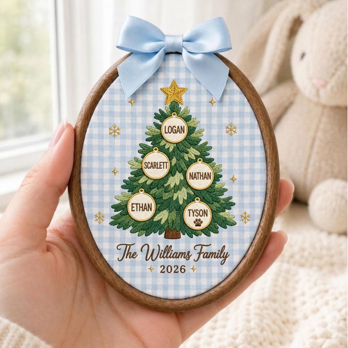 May include: An oval wooden embroidery hoop containing fabric with a light blue gingham pattern. It features an embroidered green Christmas tree with a gold star, gold snowflakes, and five circular ornaments labeled Logan, Scarlett, Nathan, Ethan, and Tyson. The text reads The Williams Family 2026, topped with a light blue satin bow.