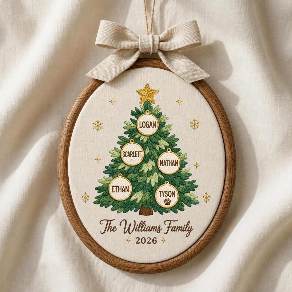 May include: An oval wooden embroidery hoop containing a cream-colored fabric with a stitched green Christmas tree. The tree features five circular ornaments with the names Logan, Scarlett, Nathan, Ethan, and Tyson, plus a paw print. Text reads The Williams Family 2026, topped with a beige bow.