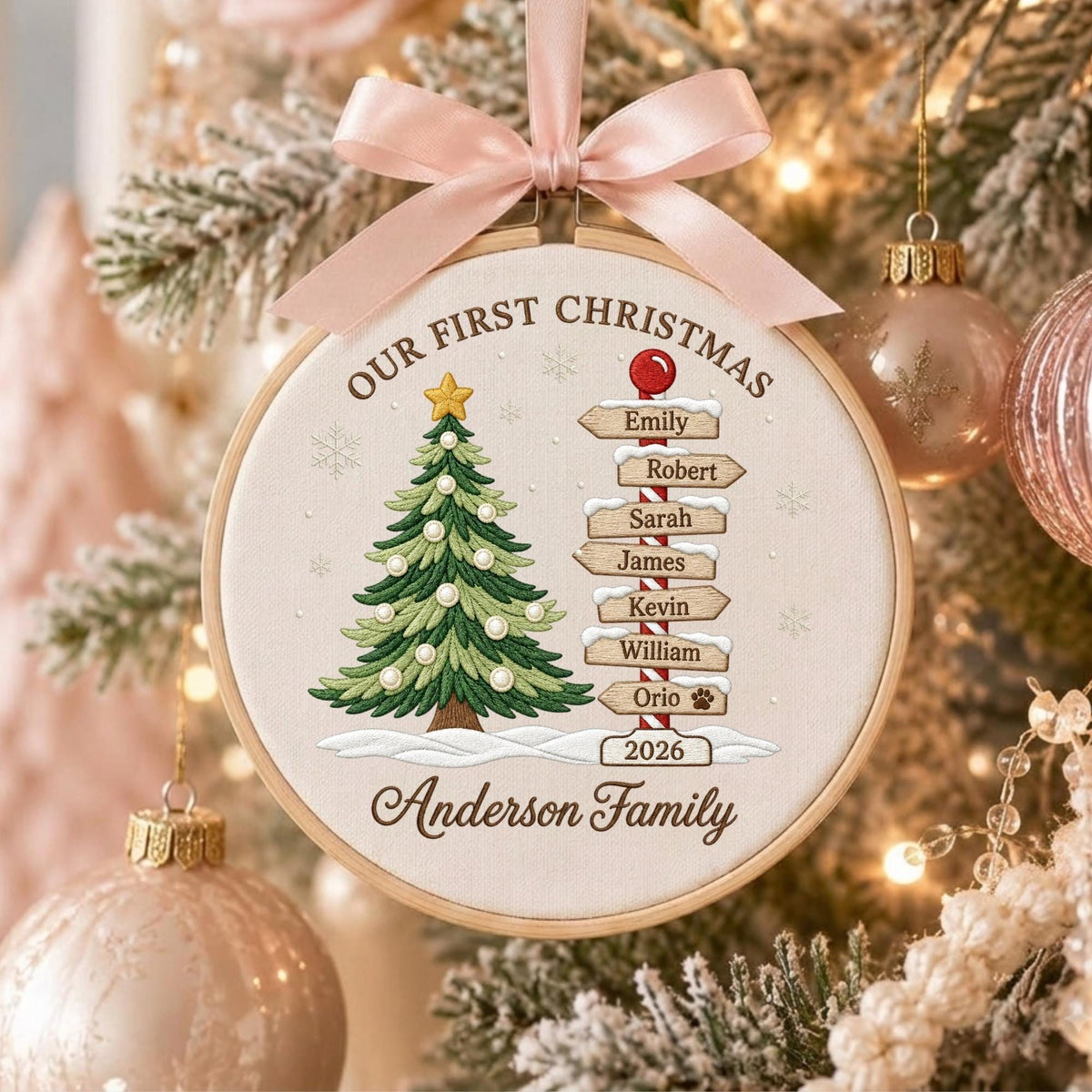 May include: A round embroidery hoop ornament featuring a light fabric background with a green Christmas tree, a red and white striped signpost with names, and the text "OUR FIRST CHRISTMAS" and "Anderson Family" with the year "2026" and a pink ribbon.