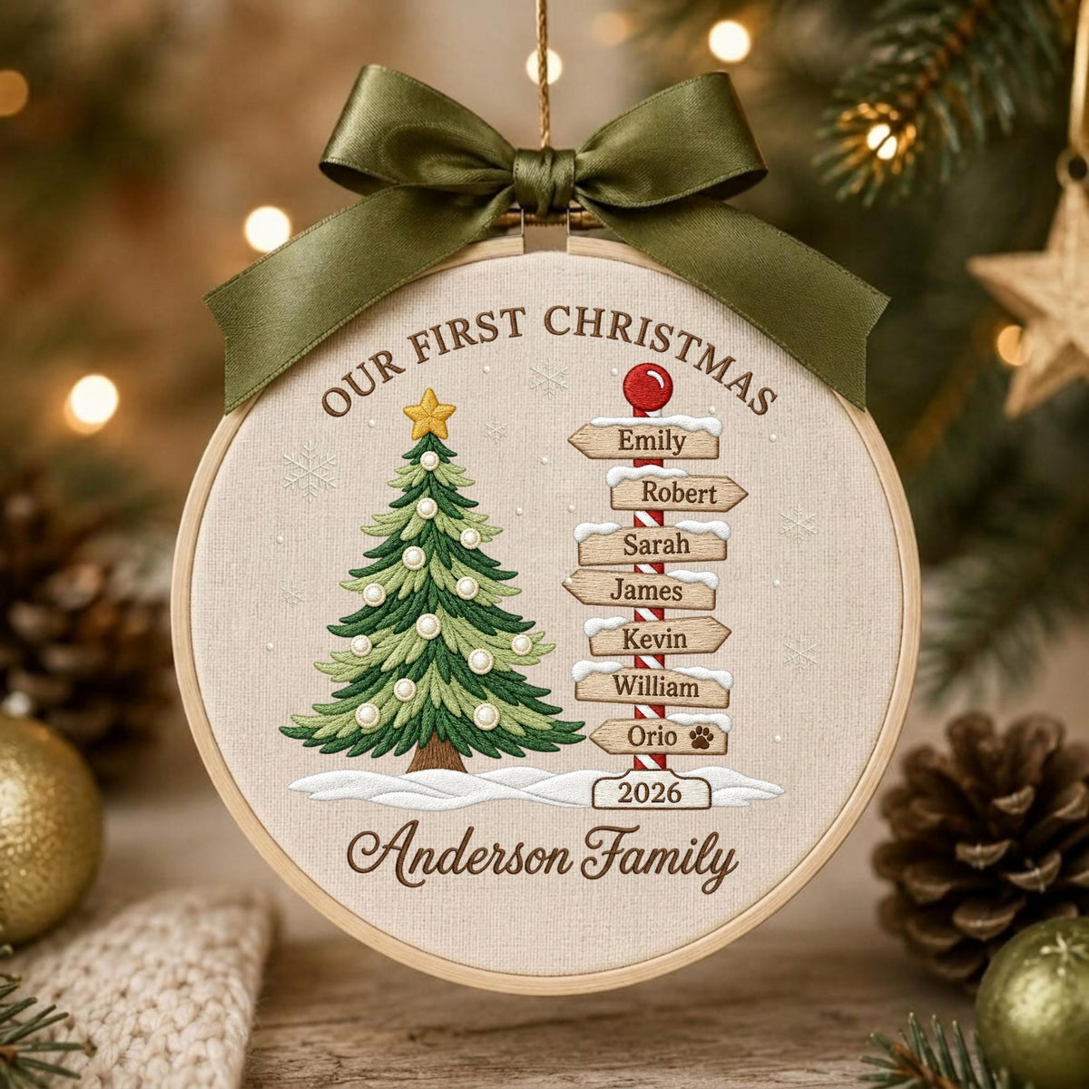 May include: A round embroidery hoop ornament featuring a beige fabric background with a green Christmas tree, a signpost with names Emily, Robert, Sarah, James, Kevin, William, Orio, and the text Our First Christmas and Anderson Family 2026.