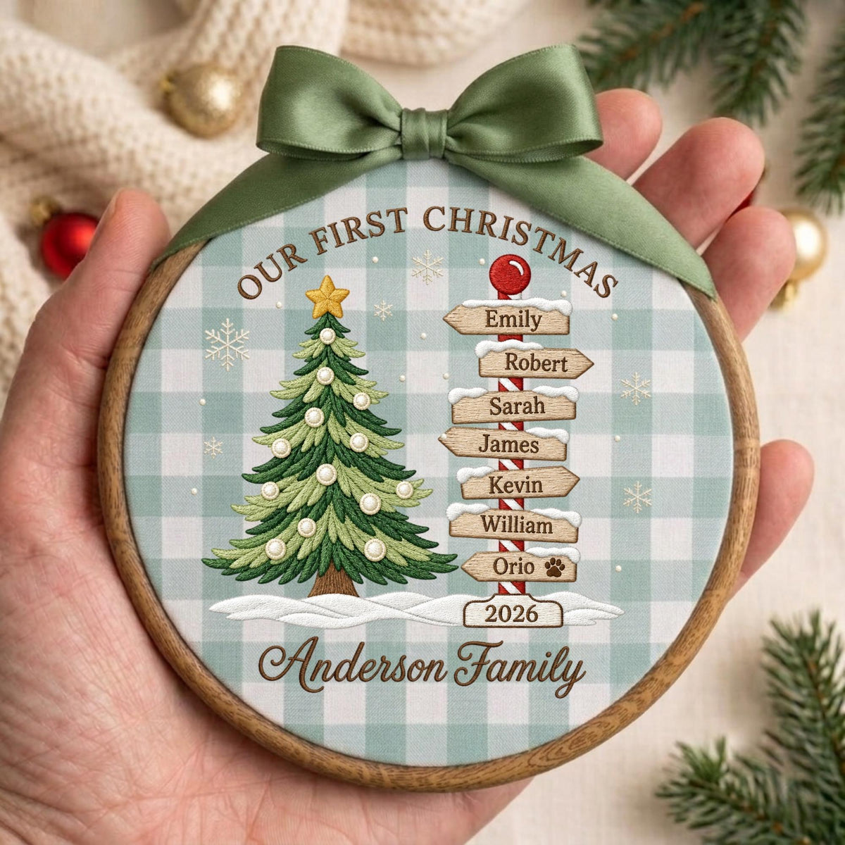 May include: A round wooden Christmas ornament featuring a green and white buffalo plaid background. It displays a green Christmas tree, a signpost with names Emily, Robert, Sarah, James, Kevin, William, Orio, and the year 2026, topped with a green ribbon.