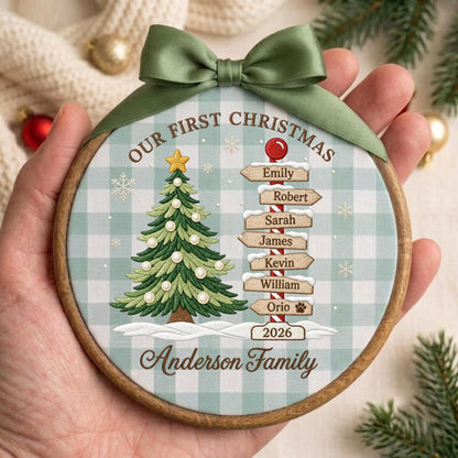 May include: A round wooden Christmas ornament featuring a green and white buffalo plaid background. It displays a green Christmas tree, a signpost with names Emily, Robert, Sarah, James, Kevin, William, Orio, and the year 2026, topped with a green ribbon.