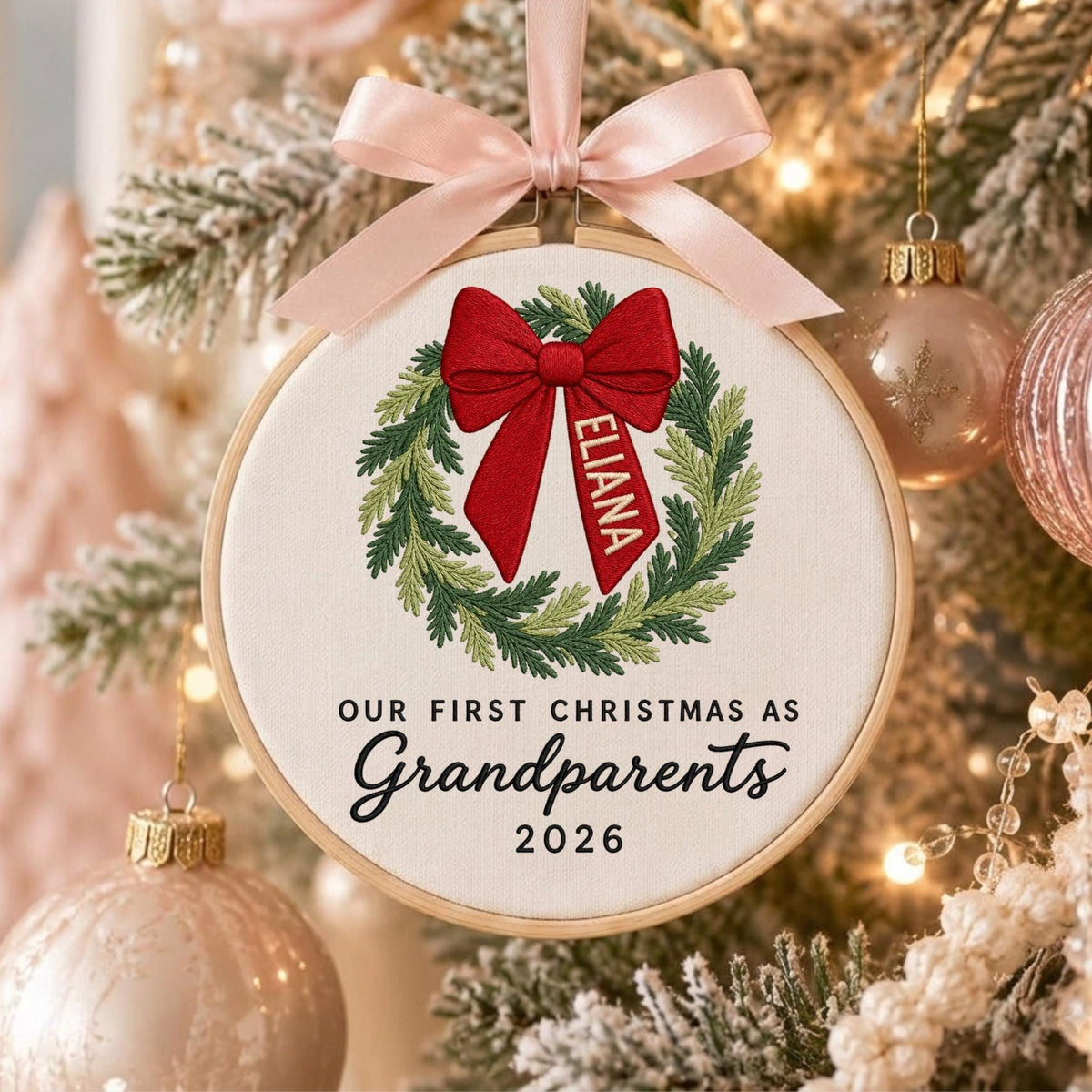 May include: A round wooden embroidery hoop ornament featuring a white fabric center with an embroidered green wreath and a large red bow. The bow has the name ELIANA printed on it. Text reads: OUR FIRST CHRISTMAS AS Grandparents 2026.