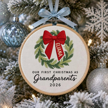 May include: A circular wooden embroidery hoop ornament featuring a stitched green wreath with a red bow. The bow has the name ELIANA embroidered in white. Below the wreath, the text reads OUR FIRST CHRISTMAS AS Grandparents 2026.