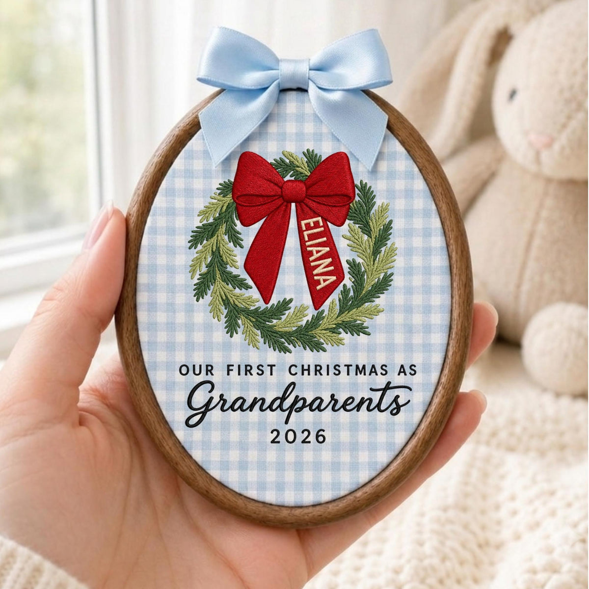 May include: An oval wooden embroidery hoop ornament featuring a light blue and white gingham fabric background. It displays an embroidered green wreath with a red bow, the name ELIANA, and the text OUR FIRST CHRISTMAS AS Grandparents 2026.