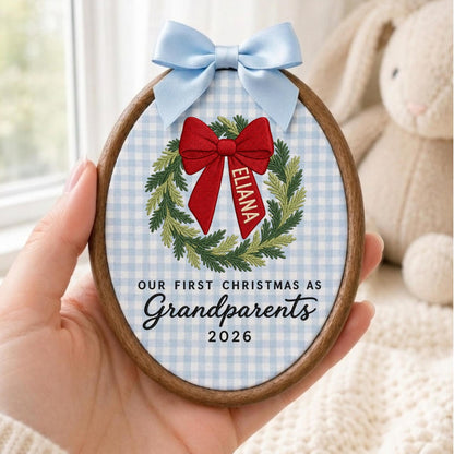 May include: An oval wooden embroidery hoop ornament featuring a light blue and white gingham fabric background. It displays an embroidered green wreath with a red bow, the name ELIANA, and the text OUR FIRST CHRISTMAS AS Grandparents 2026.