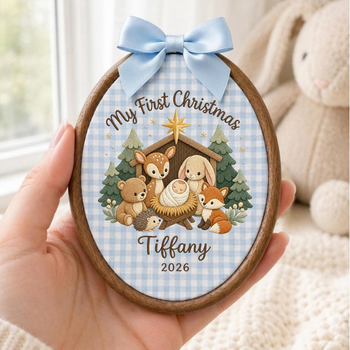 May include: An oval wooden ornament featuring a light blue gingham fabric background. The design shows a manger scene with a baby, a bear, a deer, a rabbit, a hedgehog, and a fox. Text reads My First Christmas, Tiffany, and 2026. A light blue satin bow is attached to the top.