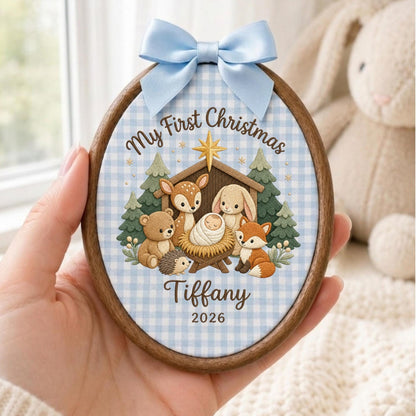 May include: An oval wooden ornament featuring a light blue gingham fabric background. The design shows a manger scene with a baby, a bear, a deer, a rabbit, a hedgehog, and a fox. Text reads My First Christmas, Tiffany, and 2026. A light blue satin bow is attached to the top.