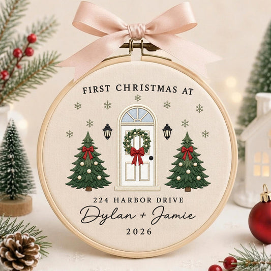 May include: A round wooden embroidery hoop containing cream fabric with a Christmas-themed design. The embroidery features a white front door with a wreath, two green Christmas trees, snowflakes, and the text "FIRST CHRISTMAS AT 224 HARBOR DRIVE Dylan + Jamie 2026". A light pink satin ribbon is tied at the top.