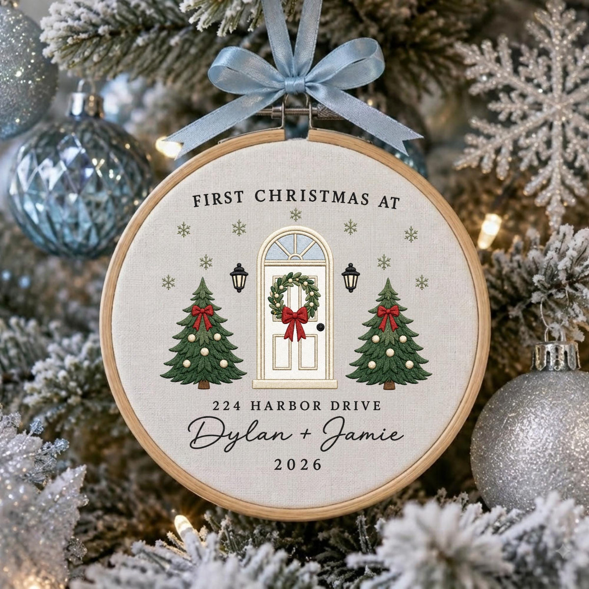 May include: A round wooden embroidery hoop ornament featuring a light fabric background with embroidered green Christmas trees, a white front door with a wreath, and the text "FIRST CHRISTMAS AT 224 HARBOR DRIVE Dylan + Jamie 2026" with a blue ribbon.