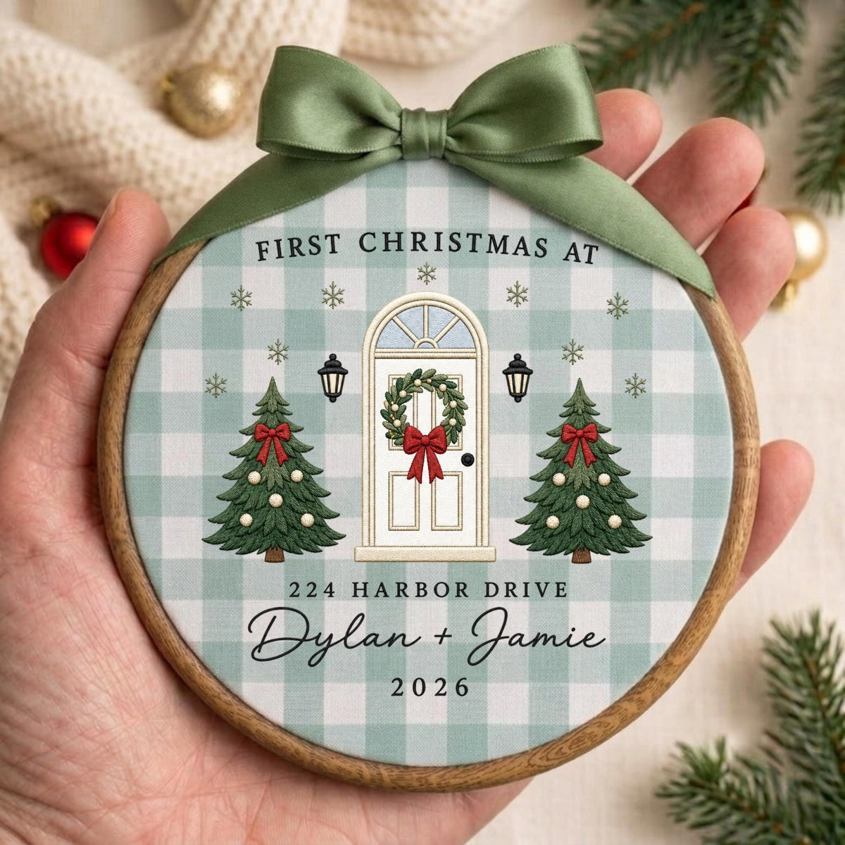 May include: A round wooden embroidery hoop ornament featuring a light green and white buffalo check fabric. The design shows a white front door with a wreath, two Christmas trees, and the text: FIRST CHRISTMAS AT 224 HARBOR DRIVE Dylan + Jamie 2026.