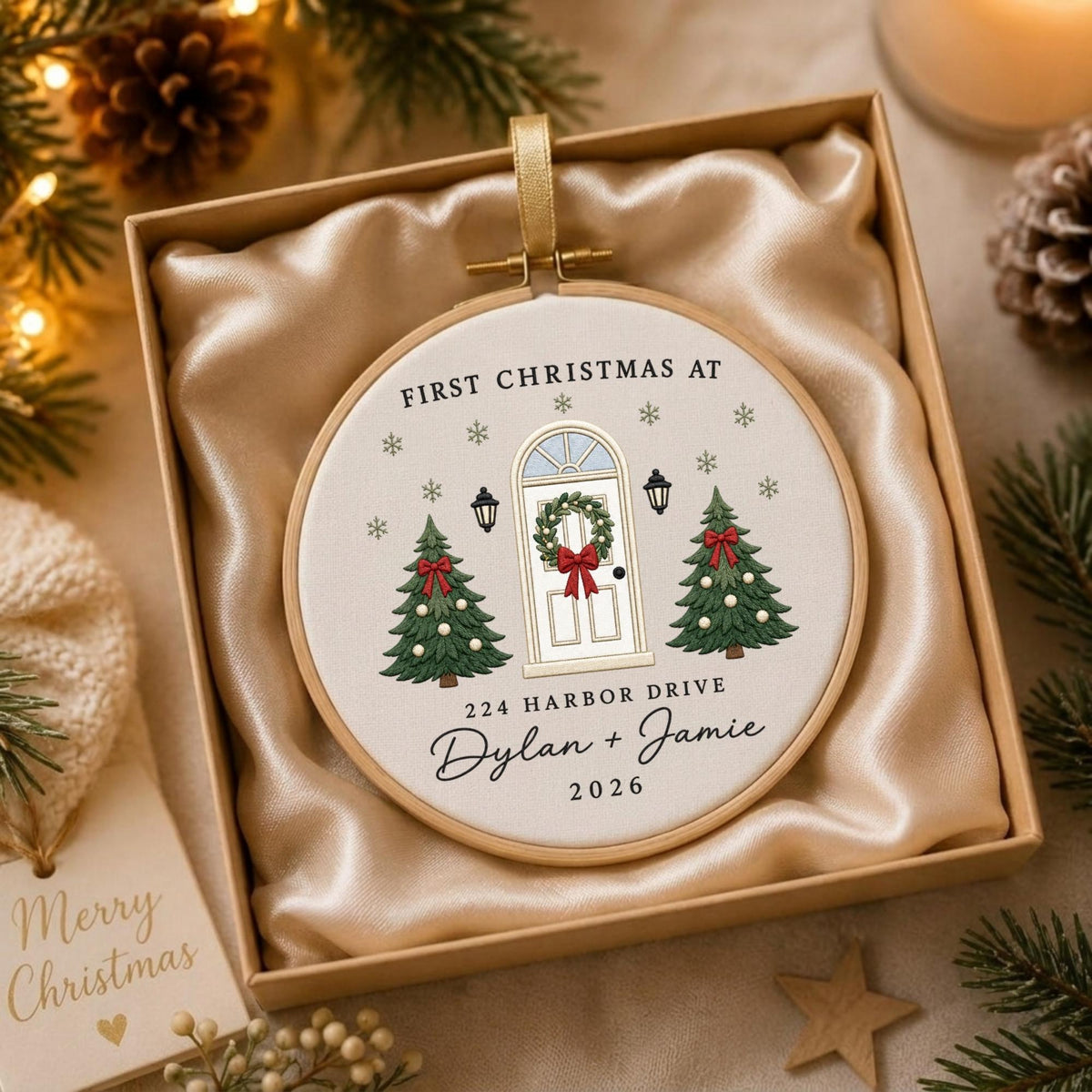 May include: A round embroidery hoop ornament featuring a white fabric center with a printed illustration of a front door flanked by two Christmas trees. The text reads FIRST CHRISTMAS AT, 224 HARBOR DRIVE, Dylan + Jamie, and 2026.