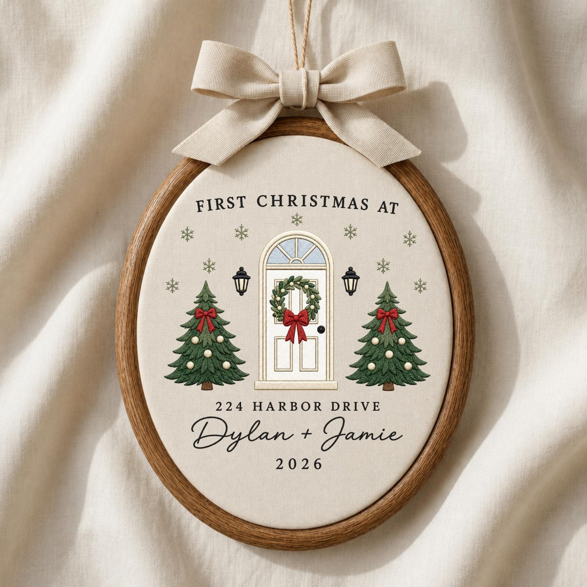 May include: An oval wooden embroidery hoop ornament featuring a cream fabric center with a printed design of a front door, two Christmas trees, and snowflakes. Text reads: FIRST CHRISTMAS AT 224 HARBOR DRIVE Dylan + Jamie 2026. Topped with a beige bow.