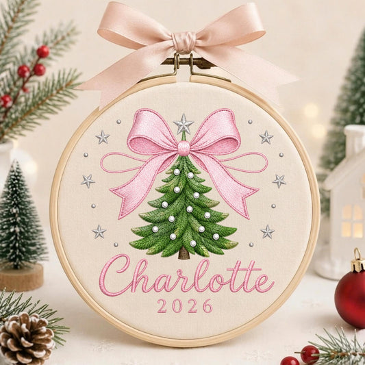 May include: A circular wooden embroidery hoop containing a cream-colored fabric panel. The fabric features an embroidered green Christmas tree with white pearl-like accents, topped with a large pink bow and a silver star. The name Charlotte and 2026 are stitched in pink below the tree. A pink satin ribbon is tied to the top of the hoop.