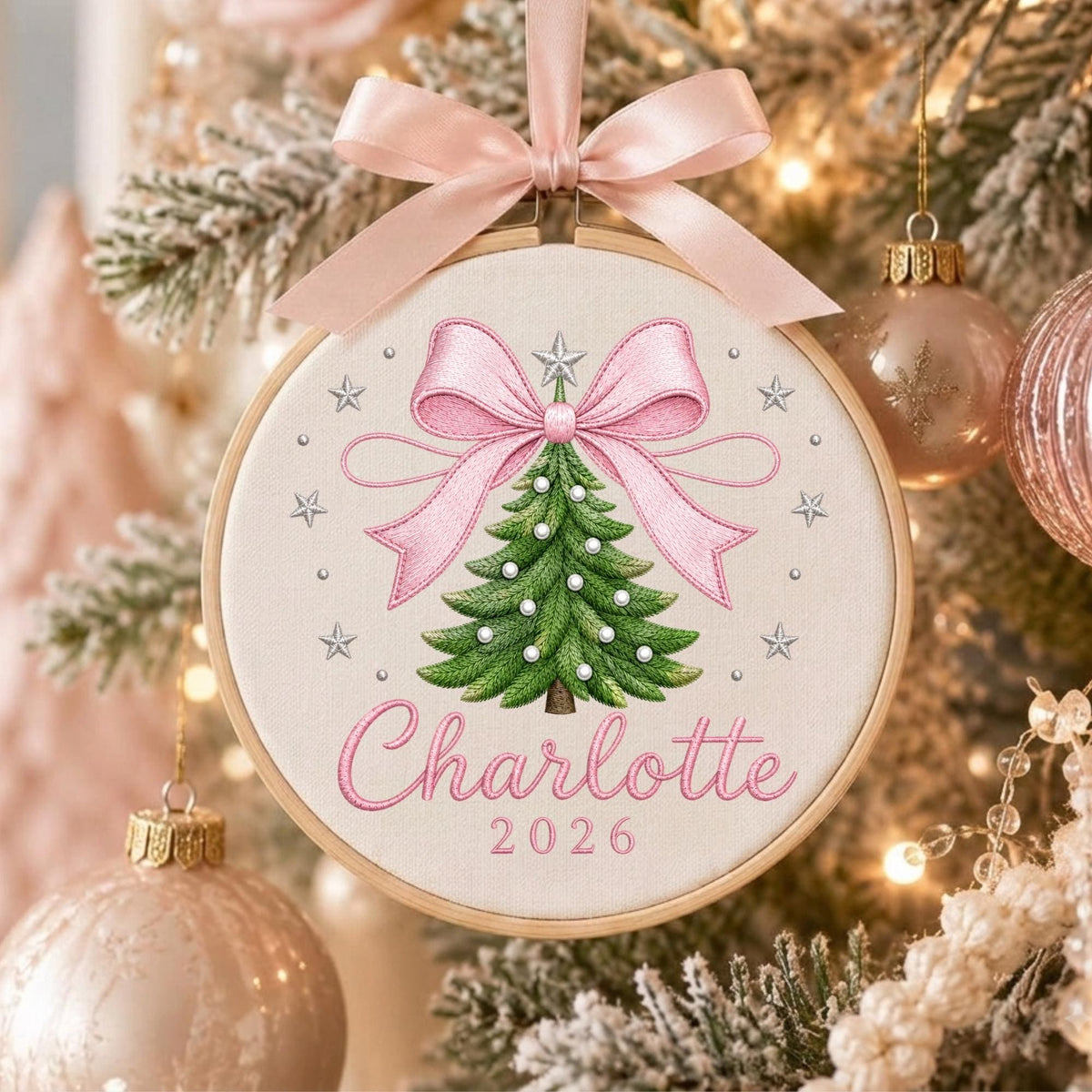 May include: A round wooden embroidery hoop ornament featuring a cream fabric center with an embroidered green Christmas tree, a large pink bow, and silver stars. The name Charlotte and the year 2026 are stitched in pink. A pink satin ribbon bow is attached.