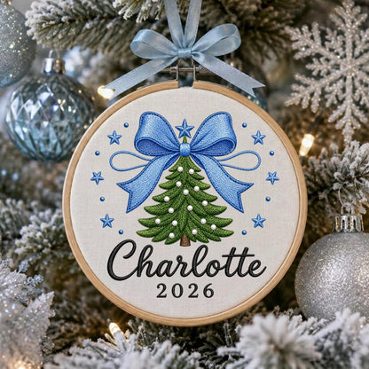 May include: A circular embroidery hoop ornament featuring a green Christmas tree with white bead accents, topped with a large blue bow and surrounded by blue stars. The name Charlotte and the year 2026 are stitched in black below the tree.