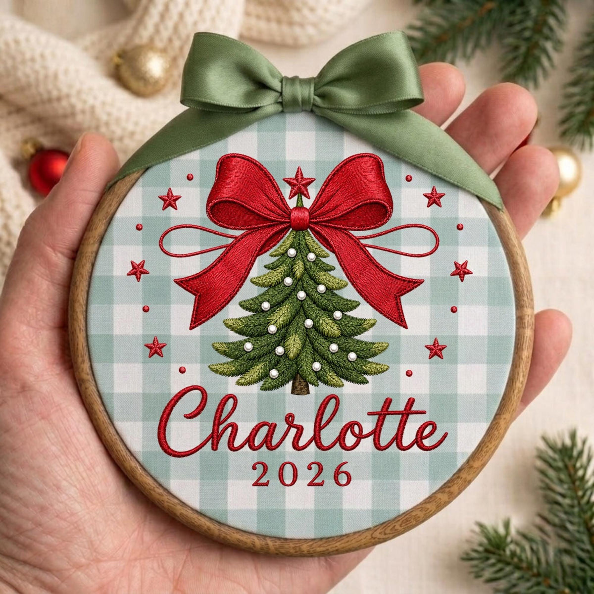 May include: A round wooden embroidery hoop ornament featuring a light green and white gingham fabric background. The design includes an embroidered green Christmas tree with white beads, a large red bow, and the red text Charlotte 2026.