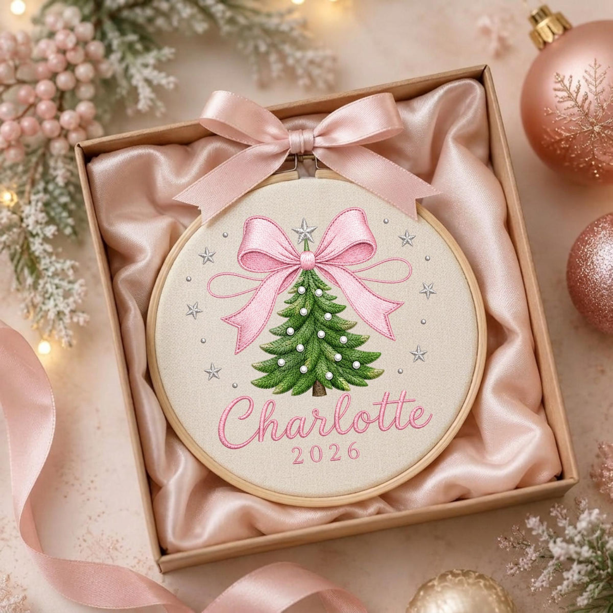 May include: A round wooden embroidery hoop containing a fabric panel with an embroidered green Christmas tree, a large pink bow, and small silver stars. The name Charlotte and the year 2026 are embroidered in pink. A pink satin ribbon is tied at the top.