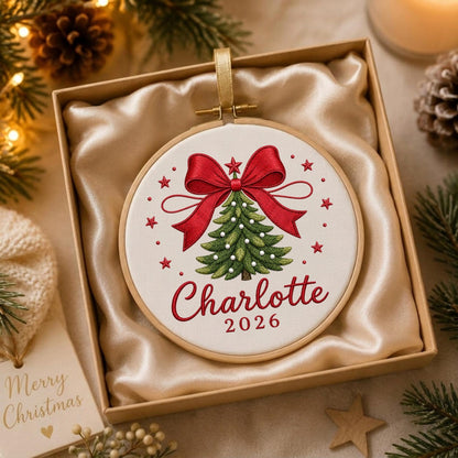 May include: A circular wooden embroidery hoop containing white fabric with an embroidered green Christmas tree, a large red bow, and small red stars. The text Charlotte and 2026 are embroidered in red script and block lettering below the tree.