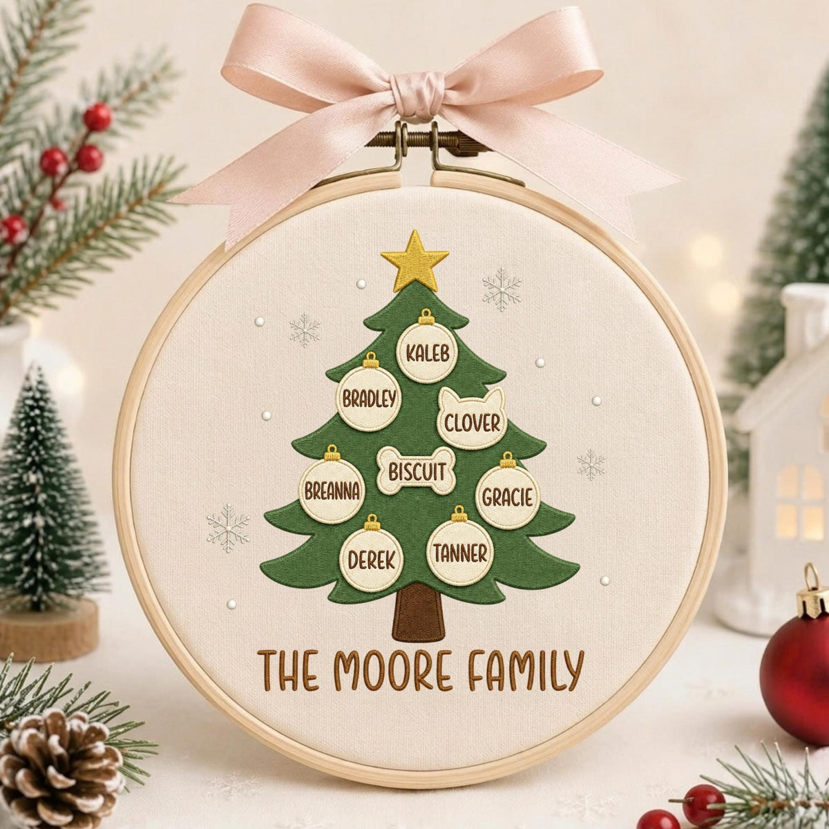 May include: A round wooden embroidery hoop containing a fabric panel with a green Christmas tree design. The tree features a gold star, white snowflake accents, and hanging ornaments labeled with names Kaleb, Bradley, Clover, Biscuit, Breanna, Gracie, Derek, and Tanner. The text THE MOORE FAMILY is embroidered at the bottom, and a light pink ribbon bow is attached to the top.