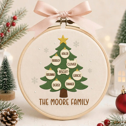 May include: A round wooden embroidery hoop containing a fabric panel with a green Christmas tree design. The tree features a gold star, white snowflake accents, and hanging ornaments labeled with names Kaleb, Bradley, Clover, Biscuit, Breanna, Gracie, Derek, and Tanner. The text THE MOORE FAMILY is embroidered at the bottom, and a light pink ribbon bow is attached to the top.