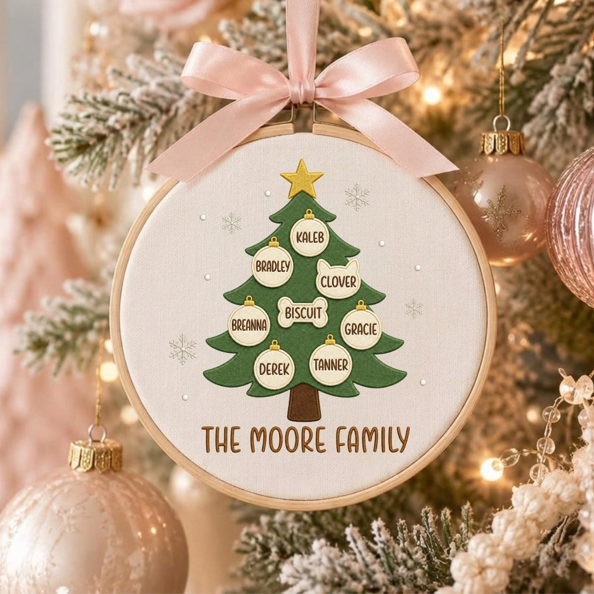 May include: A round wooden embroidery hoop ornament featuring a green Christmas tree on white fabric. The tree is topped with a gold star and decorated with white ornaments labeled Kaleb, Bradley, Clover, Biscuit, Breanna, Gracie, Derek, and Tanner. The text THE MOORE FAMILY is printed below the tree. A light pink satin ribbon bow is tied at the top.