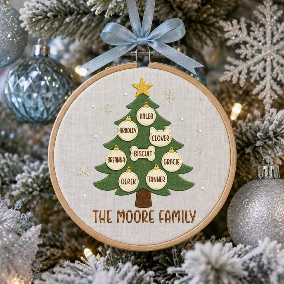 May include: A circular wooden embroidery hoop containing a fabric panel with a green Christmas tree design. The tree features a yellow star topper and several white ornaments labeled with the names Kaleb, Bradley, Clover, Biscuit, Breanna, Gracie, Derek, and Tanner. The text THE MOORE FAMILY is embroidered at the bottom, and a light blue ribbon is tied at the top.