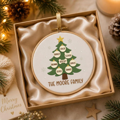 May include: A round wooden embroidery hoop ornament featuring a green Christmas tree design on white fabric. The tree is decorated with white ornaments labeled with the names Kaleb, Bradley, Clover, Biscuit, Breanna, Gracie, Derek, and Tanner. The text THE MOORE FAMILY is printed below the tree.