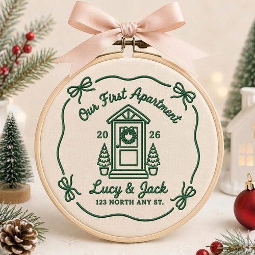 May include: A circular wooden embroidery hoop containing cream-colored fabric with dark green embroidery. The design features a house door, the text "Our First Apartment", "2026", "Lucy &amp; Jack", and "123 NORTH ANY ST.", topped with a pale pink ribbon.