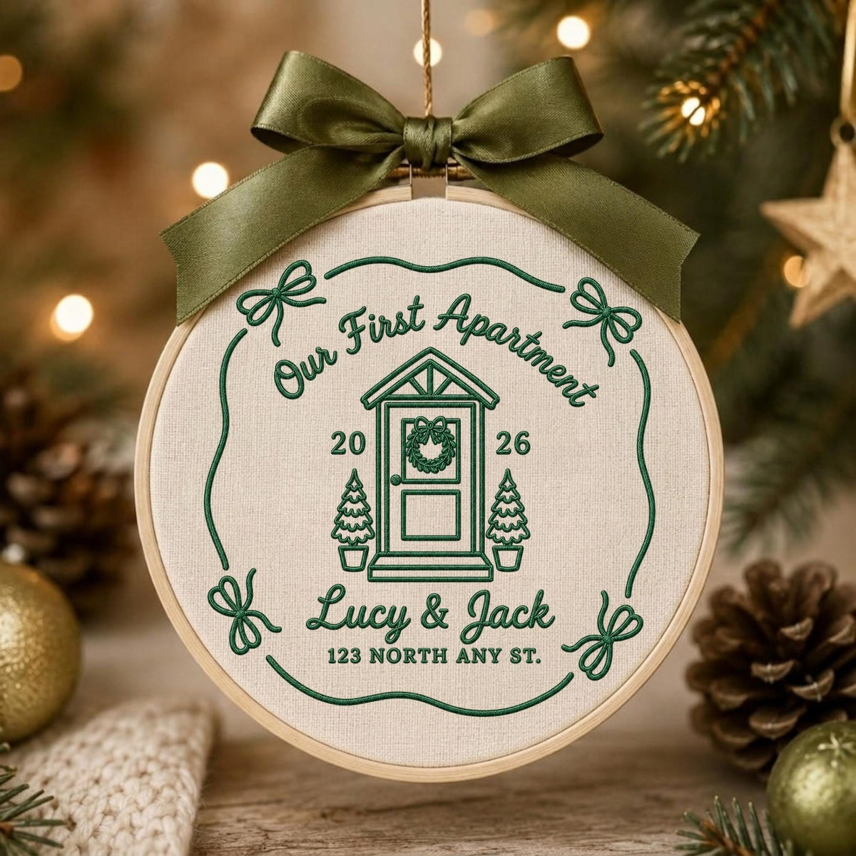 May include: A round wooden embroidery hoop containing cream-colored fabric with dark green embroidery. The design features a house icon, the text "Our First Apartment", "2026", "Lucy &amp; Jack", and "123 NORTH ANY ST.", topped with a dark green satin bow.