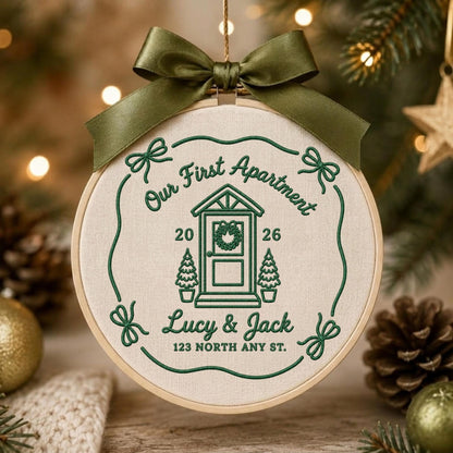 May include: A round wooden embroidery hoop containing cream-colored fabric with dark green embroidery. The design features a house icon, the text "Our First Apartment", "2026", "Lucy &amp; Jack", and "123 NORTH ANY ST.", topped with a dark green satin bow.