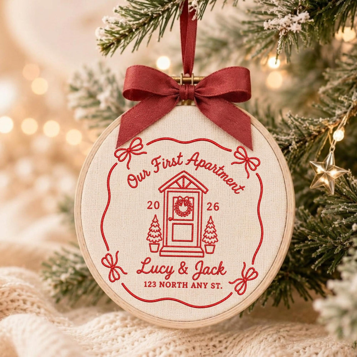 May include: A round embroidery hoop ornament featuring cream fabric with red stitched text and graphics. The design includes a house door, the text "Our First Apartment", "2026", "Lucy &amp; Jack", and "123 NORTH ANY ST." with a red ribbon bow.