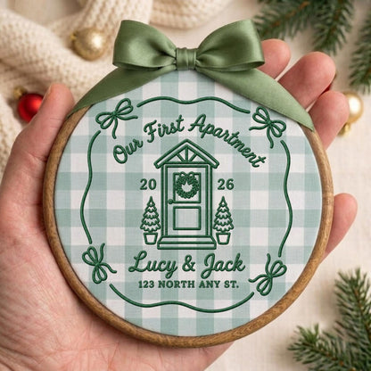 May include: A round wooden embroidery hoop ornament featuring a light green and white gingham fabric background. The center displays dark green embroidery of a house door, the text Our First Apartment, 2026, Lucy &amp; Jack, and 123 NORTH ANY ST. A sage green satin ribbon bow is tied at the top.