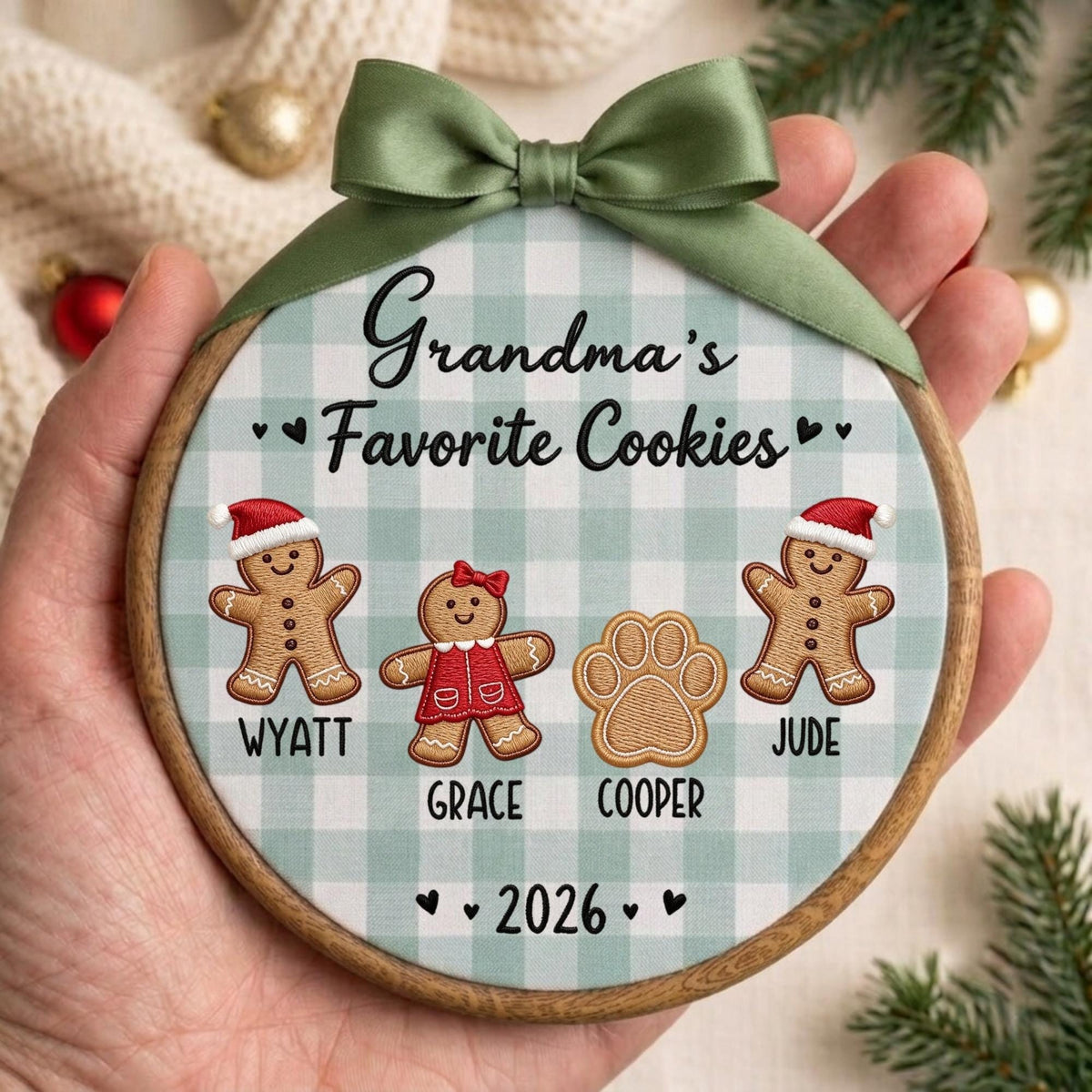 May include: A round wooden embroidery hoop ornament featuring a light green and white buffalo check fabric background. It displays the text Grandma's Favorite Cookies, gingerbread figures named Wyatt and Jude, a girl gingerbread, a paw print named Cooper, and 2026.