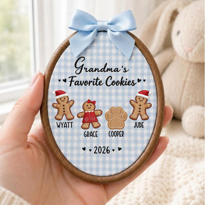 May include: An oval wooden embroidery hoop ornament featuring a light blue gingham fabric background. It displays the embroidered text "Grandma's Favorite Cookies" above three gingerbread figures and a paw print, labeled Wyatt, Grace, Cooper, and Jude, with the year 2026 and a light blue satin bow.