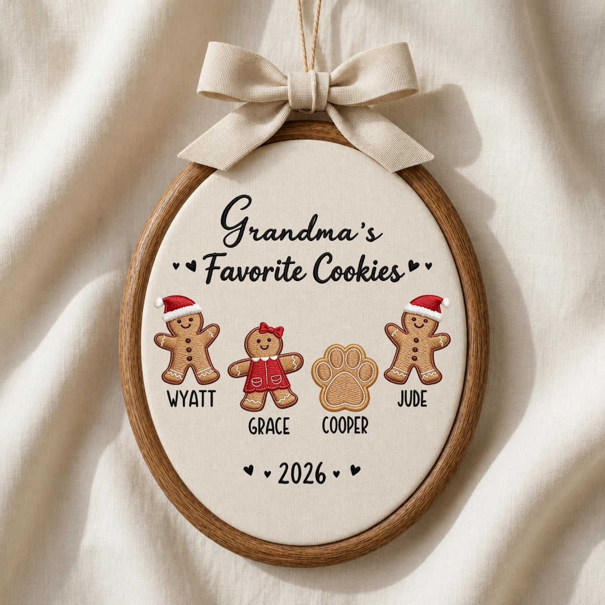 May include: An oval wooden embroidery hoop ornament featuring cream fabric with the text Grandma's Favorite Cookies. It displays two gingerbread figures in Santa hats, one gingerbread figure in a red dress, and a paw print, with names Wyatt, Grace, Cooper, Jude, and 2026, topped with a beige ribbon bow.
