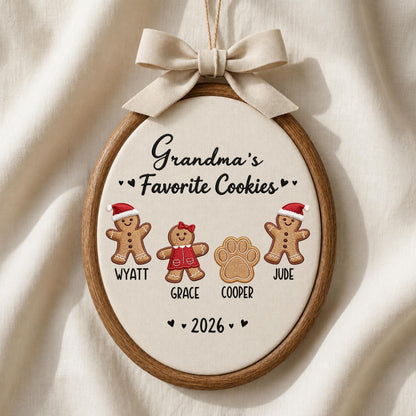 May include: An oval wooden embroidery hoop ornament featuring cream fabric with the text Grandma's Favorite Cookies. It displays two gingerbread figures in Santa hats, one gingerbread figure in a red dress, and a paw print, with names Wyatt, Grace, Cooper, Jude, and 2026, topped with a beige ribbon bow.