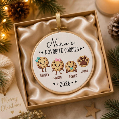 May include: A round wooden embroidery hoop ornament featuring white fabric with the text Nana's FAVORITE COOKIES and 2026. The design includes three illustrated chocolate chip cookies with faces and names Blakely, Harper, Robert, and a paw print labeled Loona.