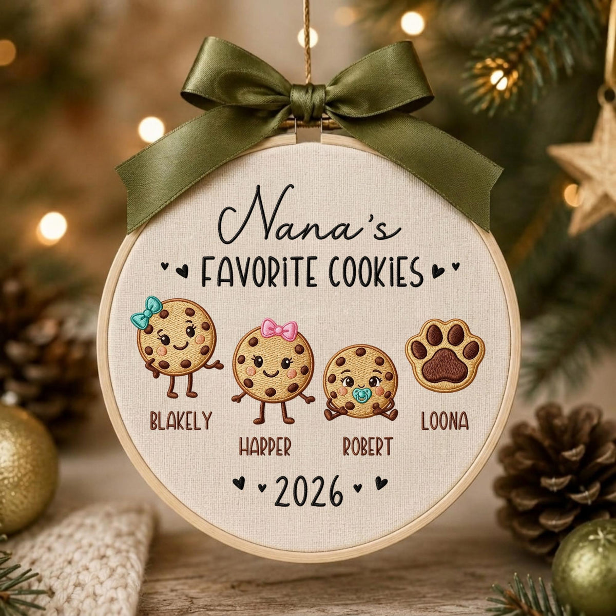 May include: A round embroidery hoop ornament featuring the text Nana's FAVORITE COOKIES and 2026. It displays three embroidered cookie characters with names Blakely, Harper, and Robert, plus a paw print labeled Loona, topped with a dark olive green ribbon.