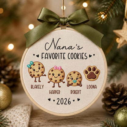 May include: A round embroidery hoop ornament featuring the text Nana's FAVORITE COOKIES and 2026. It displays three embroidered cookie characters with names Blakely, Harper, and Robert, plus a paw print labeled Loona, topped with a dark olive green ribbon.
