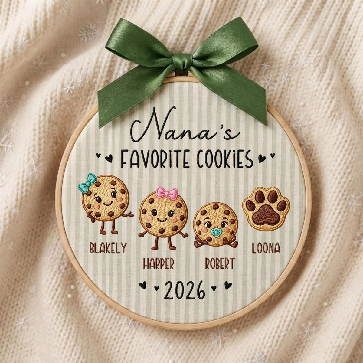May include: A round wooden embroidery hoop featuring light green and white vertical stripes. The text reads Nana's FAVORITE COOKIES, with embroidered illustrations of three cookies and a paw print, labeled Blakely, Harper, Robert, and Loona, plus 2026.