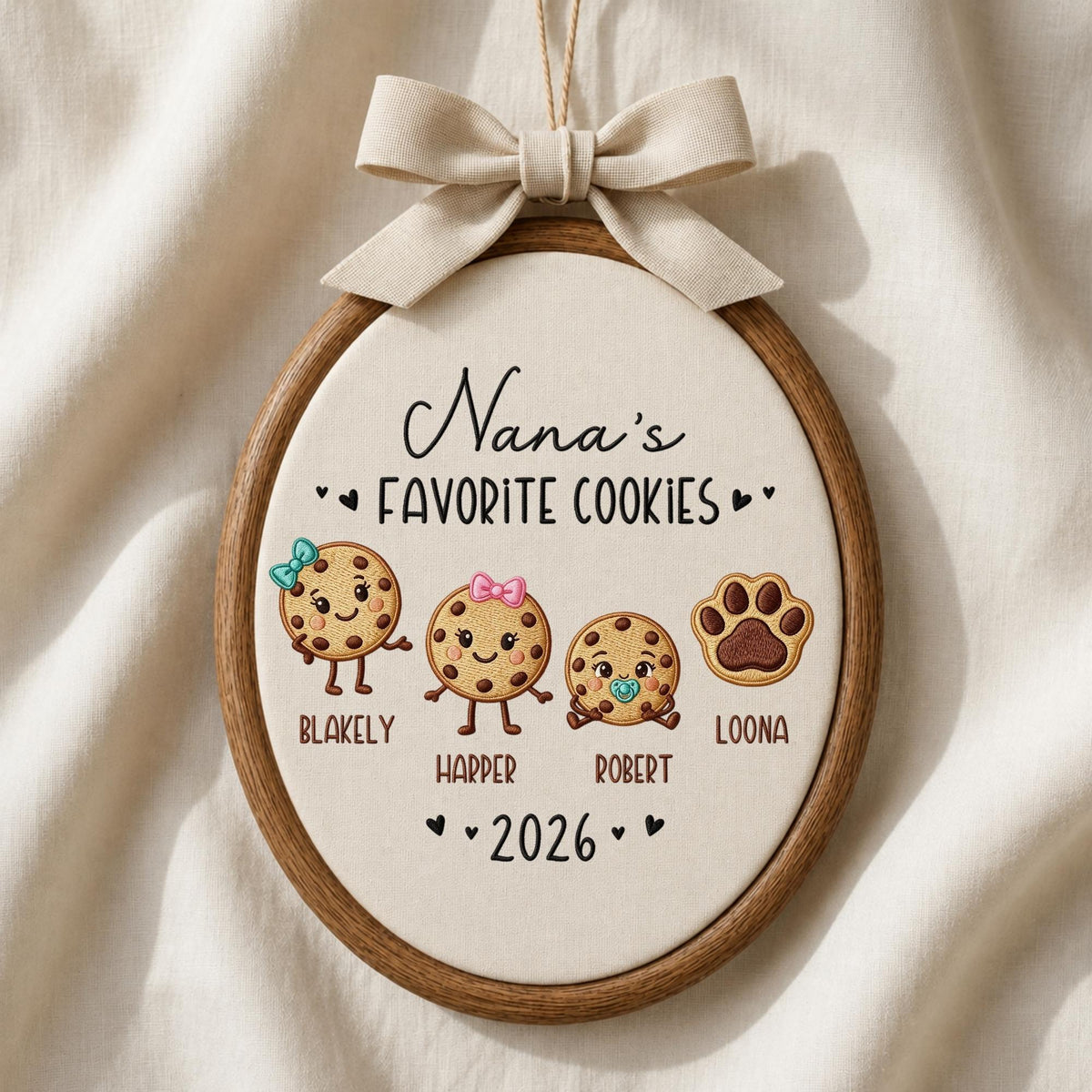 May include: An oval embroidery hoop wall hanging featuring the text Nana's FAVORITE COOKIES and 2026. It displays three cartoon cookies with faces and names Blakely, Harper, and Robert, plus a brown paw print labeled Loona, topped with a beige bow.