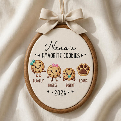 May include: An oval embroidery hoop wall hanging featuring the text Nana's FAVORITE COOKIES and 2026. It displays three cartoon cookies with faces and names Blakely, Harper, and Robert, plus a brown paw print labeled Loona, topped with a beige bow.