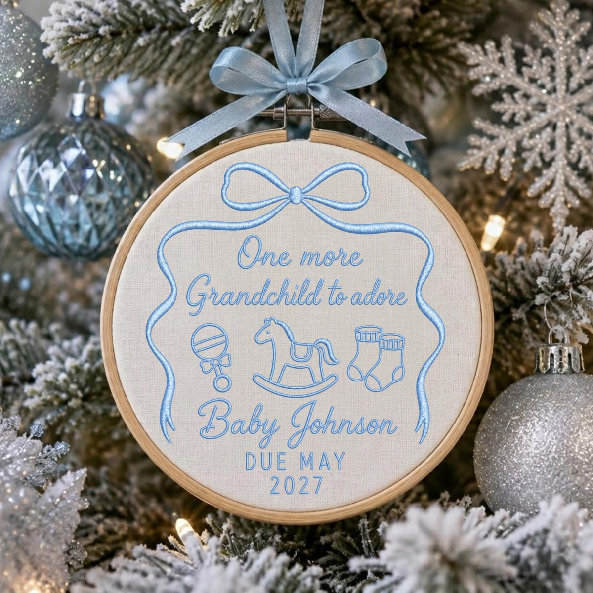 May include: A round wooden embroidery hoop ornament featuring light blue stitching on a cream fabric background. The text reads One more Grandchild to adore, Baby Johnson, DUE MAY 2027, with embroidered icons of a rattle, rocking horse, and booties.
