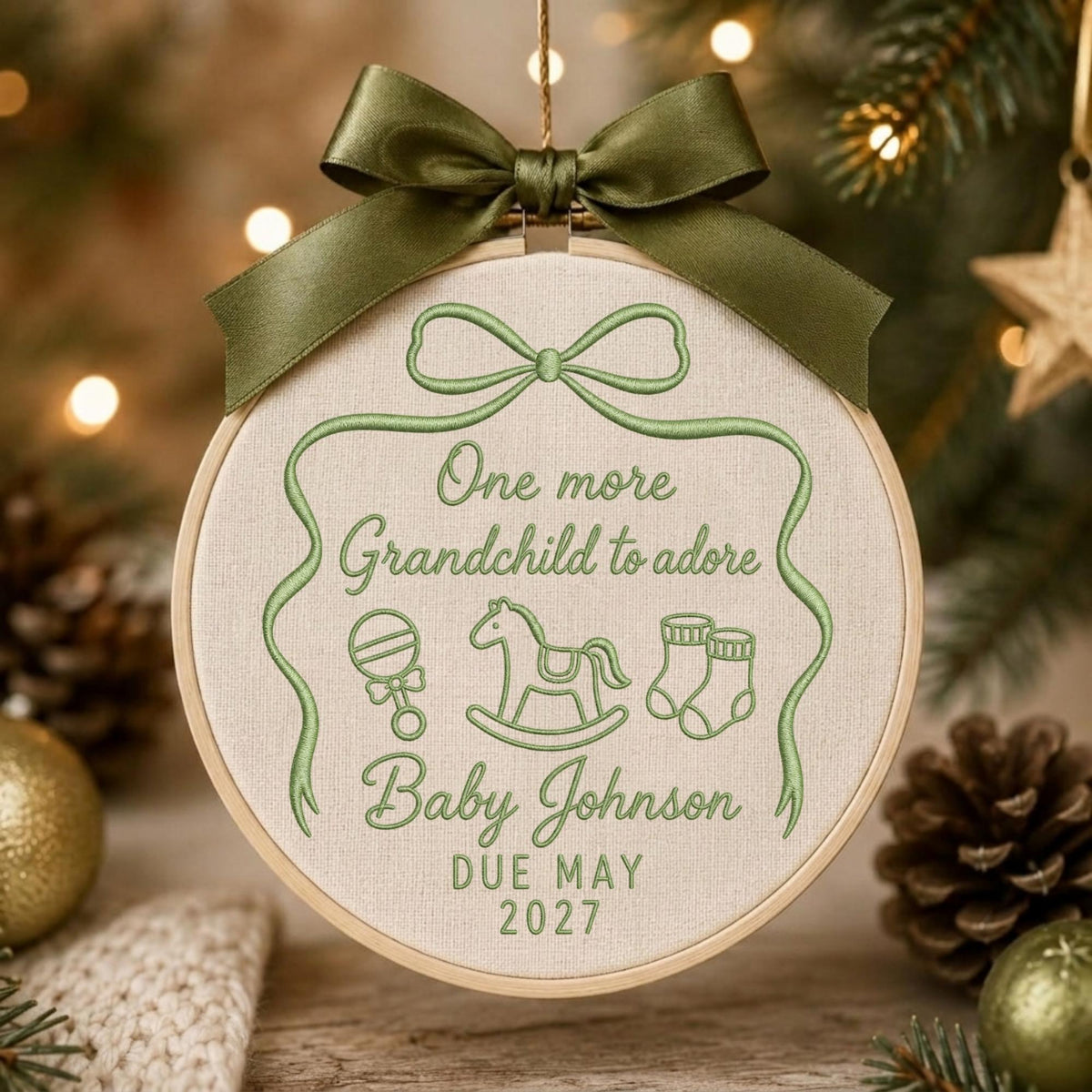 May include: A round wooden embroidery hoop ornament featuring light green thread embroidery on cream fabric. The text reads One more Grandchild to adore, Baby Johnson, DUE MAY 2027, with icons of a rattle, rocking horse, and booties, topped with a dark olive green satin ribbon bow.