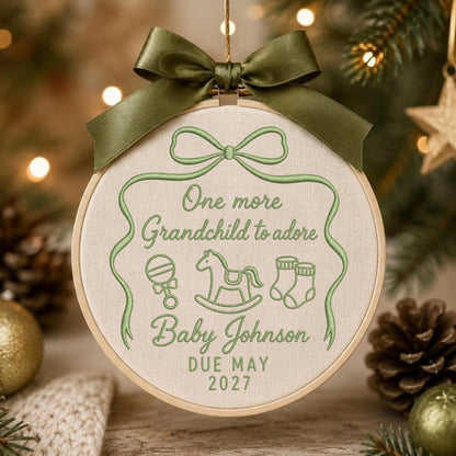 May include: A round wooden embroidery hoop ornament featuring light green thread embroidery on cream fabric. The text reads One more Grandchild to adore, Baby Johnson, DUE MAY 2027, with icons of a rattle, rocking horse, and booties, topped with a dark olive green satin ribbon bow.