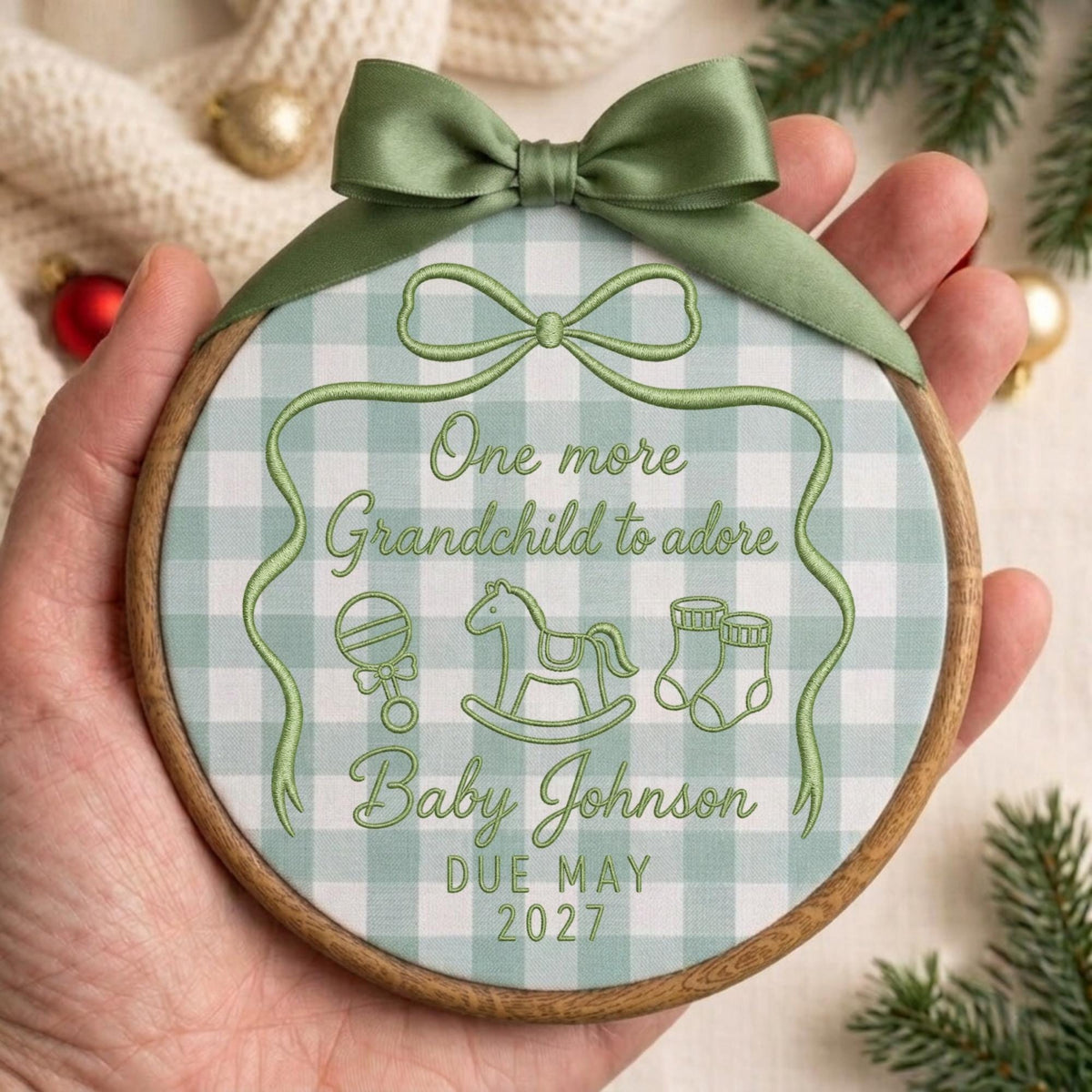May include: A round wooden embroidery hoop frame containing fabric with a sage green and white gingham pattern. The fabric features green embroidered text reading One more Grandchild to adore, Baby Johnson, and DUE MAY 2027, with icons of a rattle, rocking horse, and baby booties, topped with a sage green satin ribbon bow.