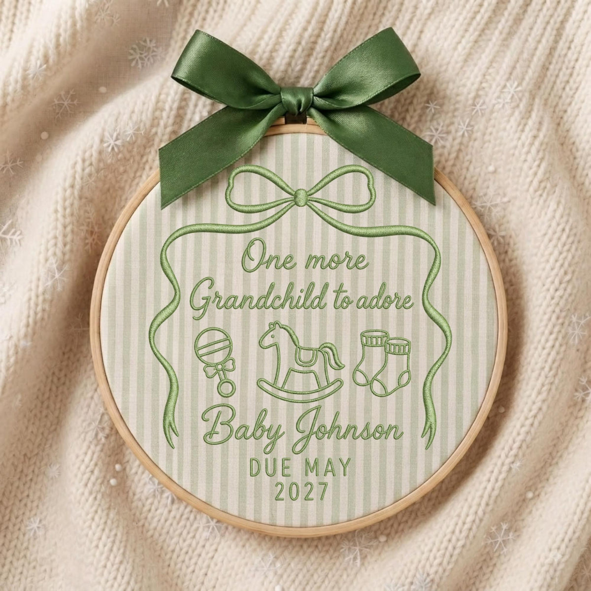 May include: A round wooden embroidery hoop containing fabric with vertical light green stripes. The fabric features green embroidery reading One more Grandchild to adore, Baby Johnson, DUE MAY 2027, with icons of a rattle, rocking horse, and socks, topped with a dark green satin bow.