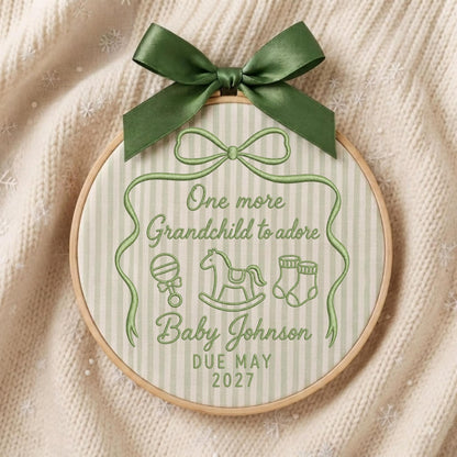 May include: A round wooden embroidery hoop containing fabric with vertical light green stripes. The fabric features green embroidery reading One more Grandchild to adore, Baby Johnson, DUE MAY 2027, with icons of a rattle, rocking horse, and socks, topped with a dark green satin bow.